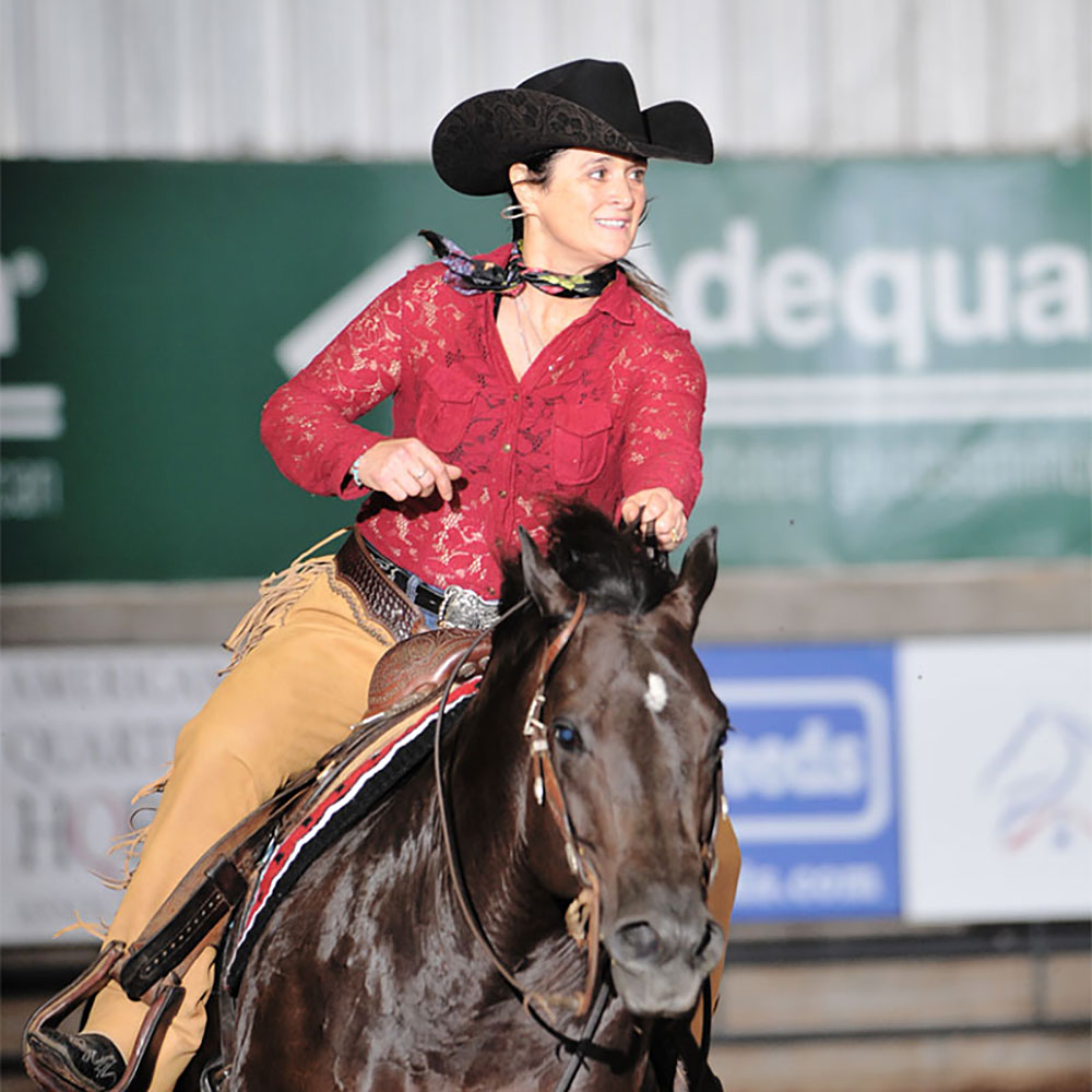 Woman in red shirt and black cowboy hat riding a dark horse in an arena.
